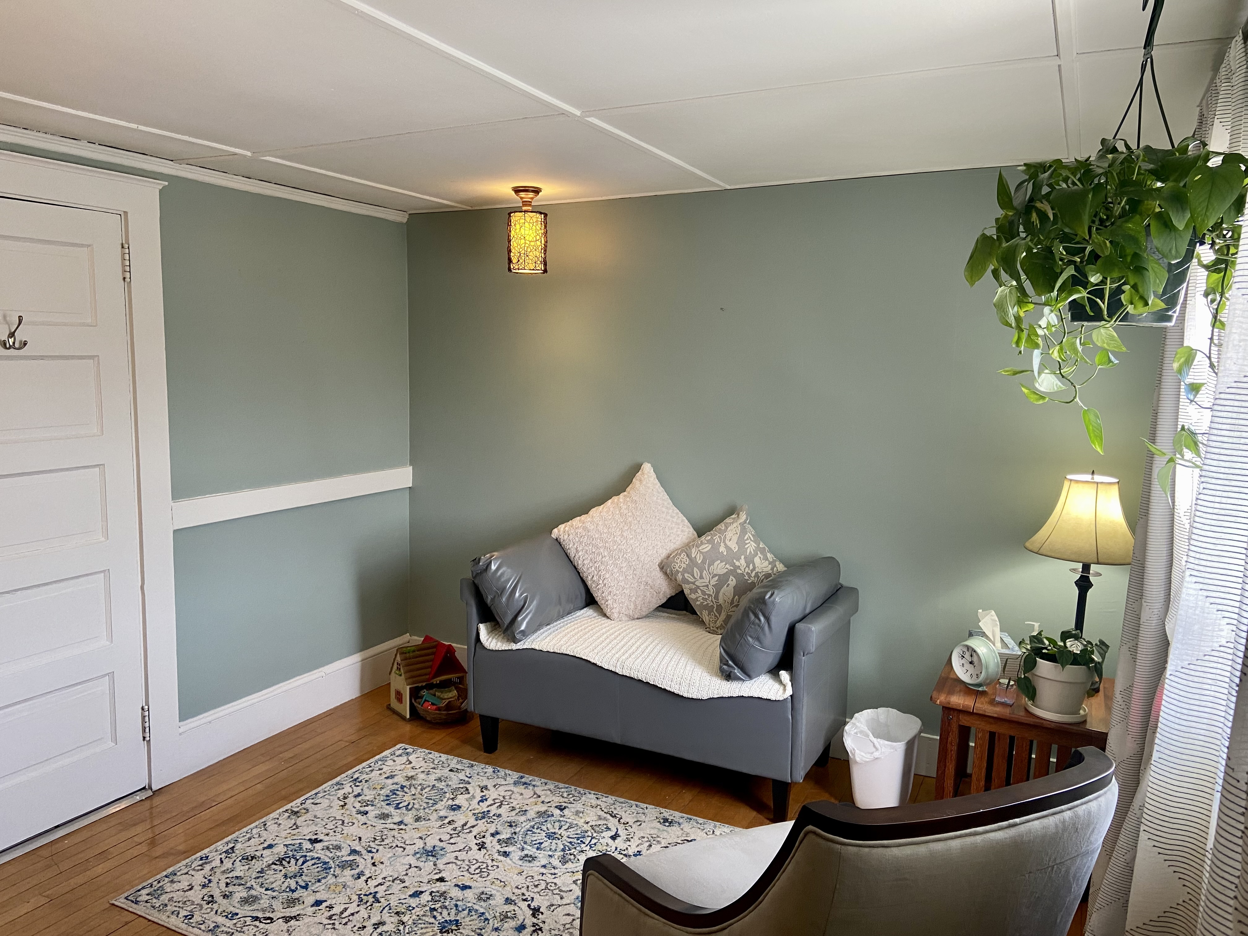 A therapist's office has a light blue wall and gray sofas on the wood floor. Green plant hanging from sealing nearby window.