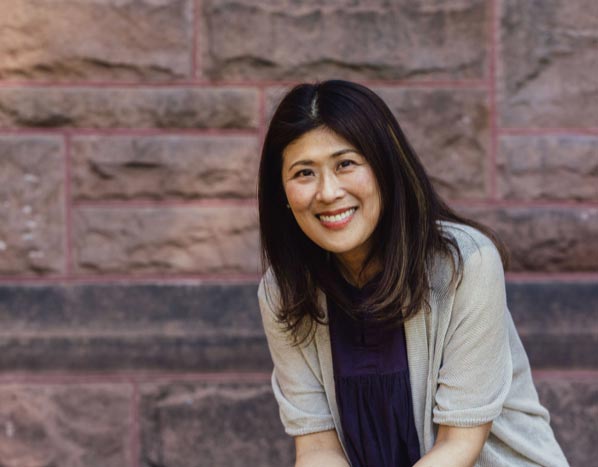 Fusako Yamagiwa smiles, looking at the camera, in front of a stone wall.